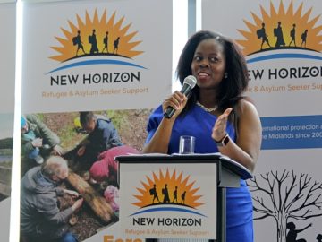 A woman wearing a blue dress gives a speech, standing behind a podium which says New Horizon on it