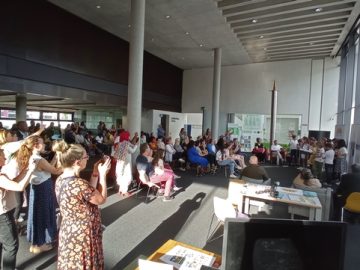 A very large group of people sit and stand in a library, clapping someone who has just given a speech