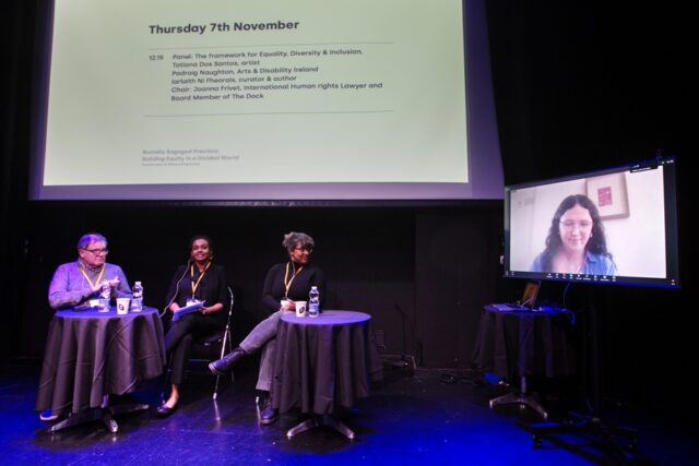 Three people sit on a stage, facing a screen on which a fourth person is appearing