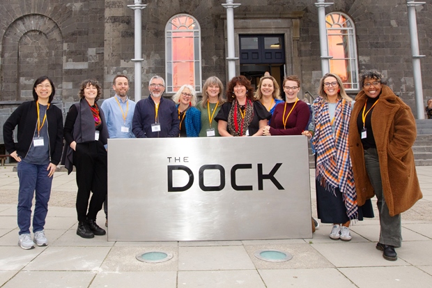 Eleven people, each wearing yellow lanyards, stand behind a large sign which says "the Dock" on it. They are smiling towards the camera