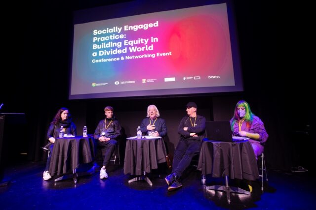 Five people sit on a stage, in front of a large screen