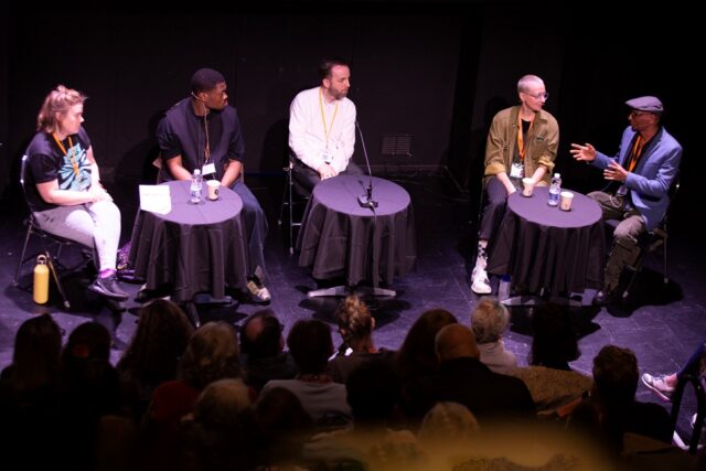 Five people sit on a stage, in front of an audience