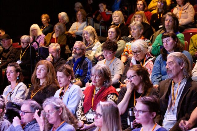 An audience of people sit in rows all looking in the same direction, towards a stage