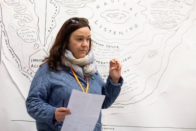 A woman with brown hear, wearing a blue jacket and a scarf , stands in front of a sketch