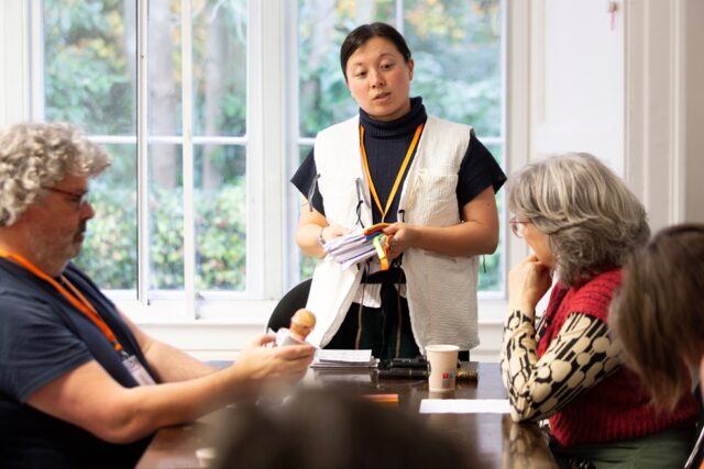 A woman wearing a black jumper and white vest stands in front of three people sitting at a table
