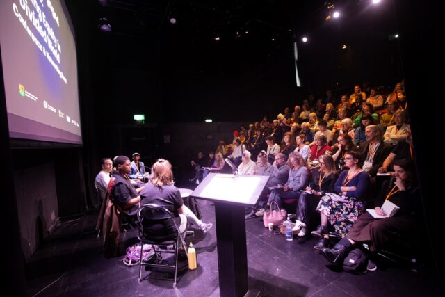 Five people sit on a stage, in front of an audience