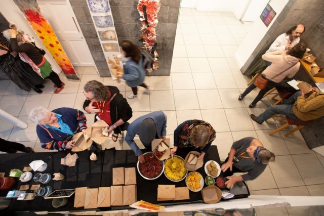 A photo taken from above shows people serving themselves food and talking, in a room with a tiled floor