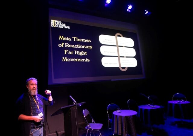 A man with a beard stands in front of a large screen which says "meme themes of reactionary far right movements" on it