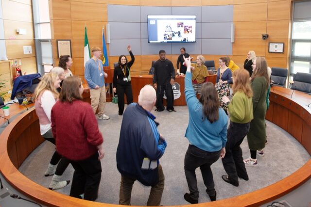 A group of people are standing inside a circular room, moving their bodies, being led by a man who stands among them