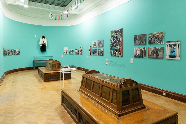 An exhibition room with large wooden seats, artworks on the walls against a turquoise background