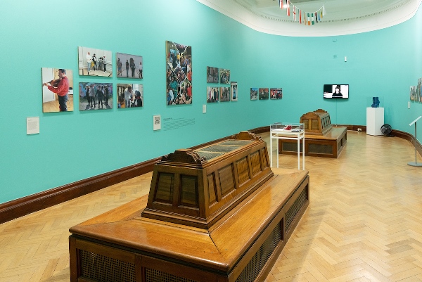 An exhibition room with large wooden seats, artworks on the walls against a turquoise background