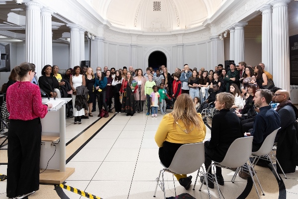 A large group of people face in one direction, watching a woman in a pink jumper make a speech