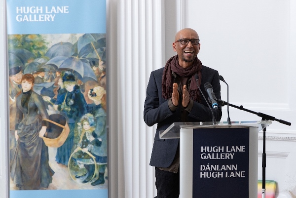 A man wearing a navy suit and brown scarf stands behind a podium which says Hugh lane Gallery on it. He is clapping and smiling