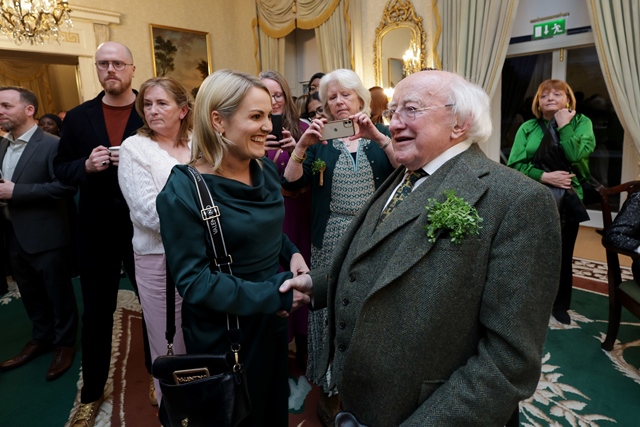 A woman shakes the hand of President of Ireland, Michael D Higgins in a large ornately decorated room. She is smiling broadly
