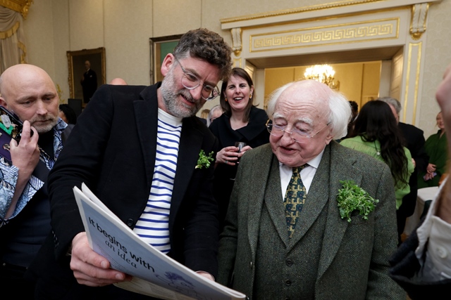 A man shows a newspaper to President of Ireland, Michael D Higgins, in a large ornately decorated room