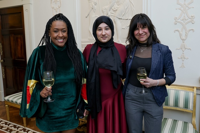 Three women stand side by side, smiling at the camera. Two of them hold wine glasses