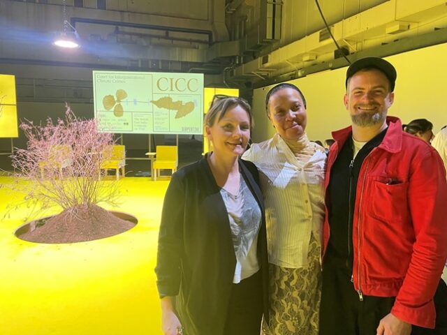 Three people smile in the foreground of the picture, with a yellow stage and a tree behind them