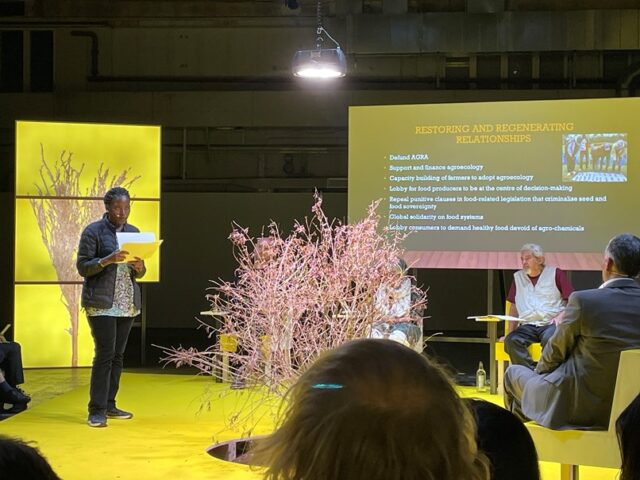 Three people sit on a yellow stage, while one person stands holding some pages and speaking to them