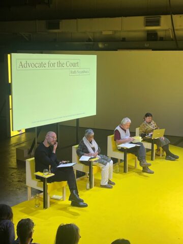 Four people sit on a yellow stage, with a screen behind them which says "advocate for the court: Ruth Nyambura" on it