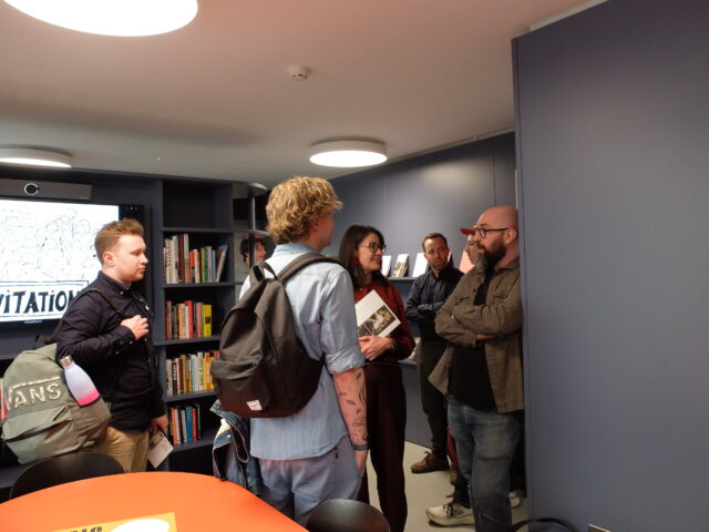 Seven people stand in a room, clustered in small groups. They are smiling and talking to each other