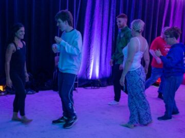 Six people move around a dark space which is dimly lit by floor lighting. They are smiling.